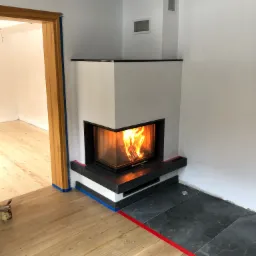 Nowoczesny kominek narożny z czarnym kamieniem i palącym się ogniem w surowym wnętrzu, zabezpieczony taśmą malarską podczas prac wykończeniowych.