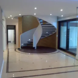 Wnętrze z spiralnymi schodami, beżową podłogą z ciemnymi akcentami i oświetleniem punktowym oraz ledowym na suficie. Ściana za schodami pokryta dekoracyjną tapetą w odcieniach złota.