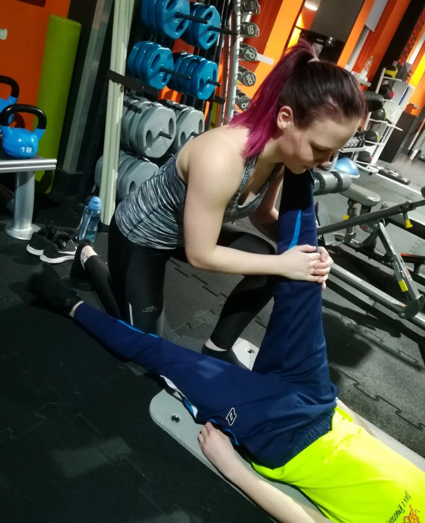 Kobieta z różowymi włosami pomaga w rozciąganiu nogi leżącemu mężczyźnie w sportowym stroju na tle siłowni z ciężarami.