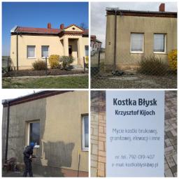 Kostka Błysk - Pracownik w niebieskiej czapce myje elewację budynku z żółtym tynkiem za pomocą myjki ciśnieniowej, widoczny efekt czyszczenia oraz tabliczka z ofertą usług: mycie kostki brukowej, elewacji.