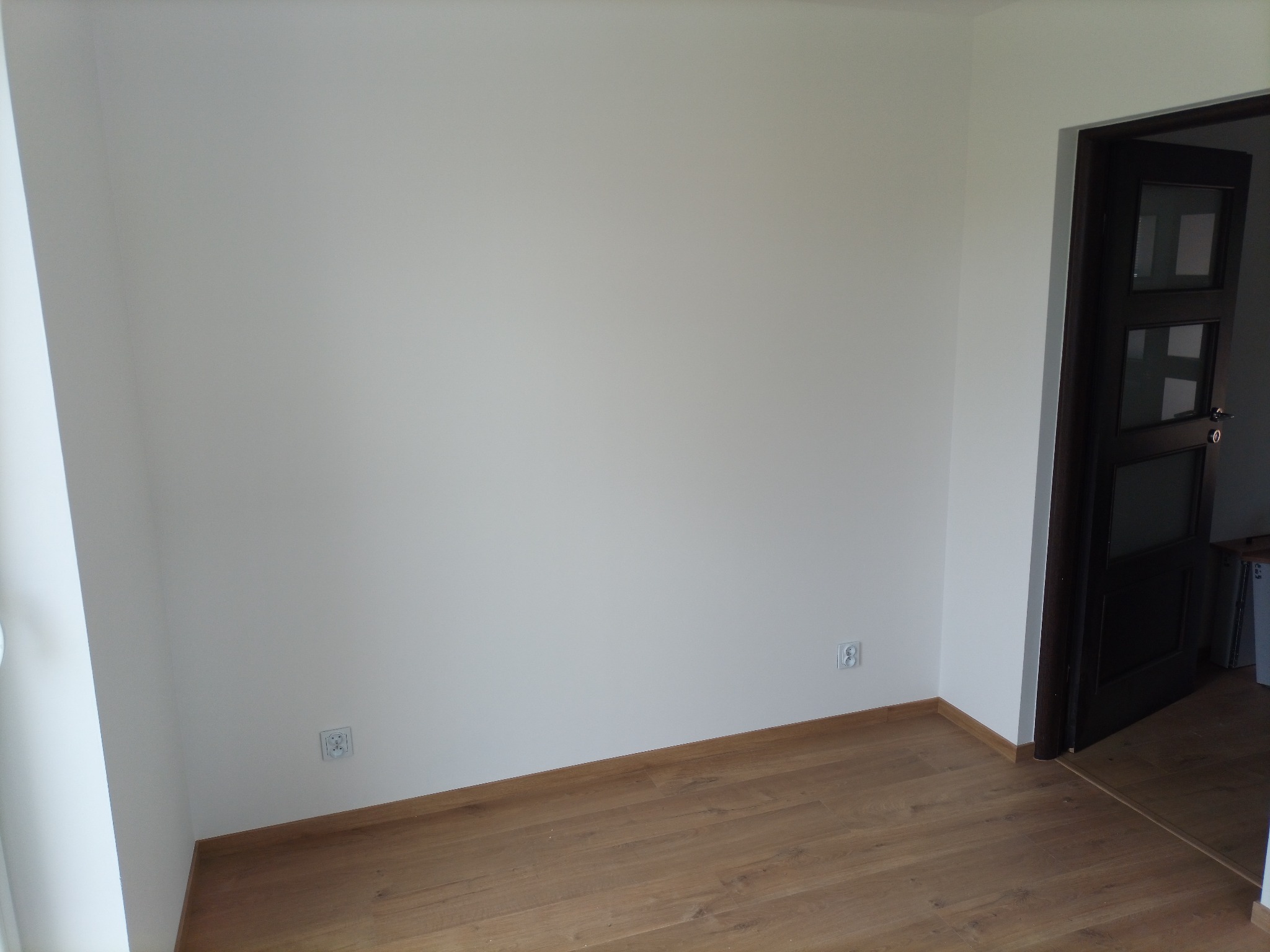 Pomalowane na biało ściany w pustym pokoju z drewnianą podłogą i ciemnymi drzwiami z przeszkleniami.