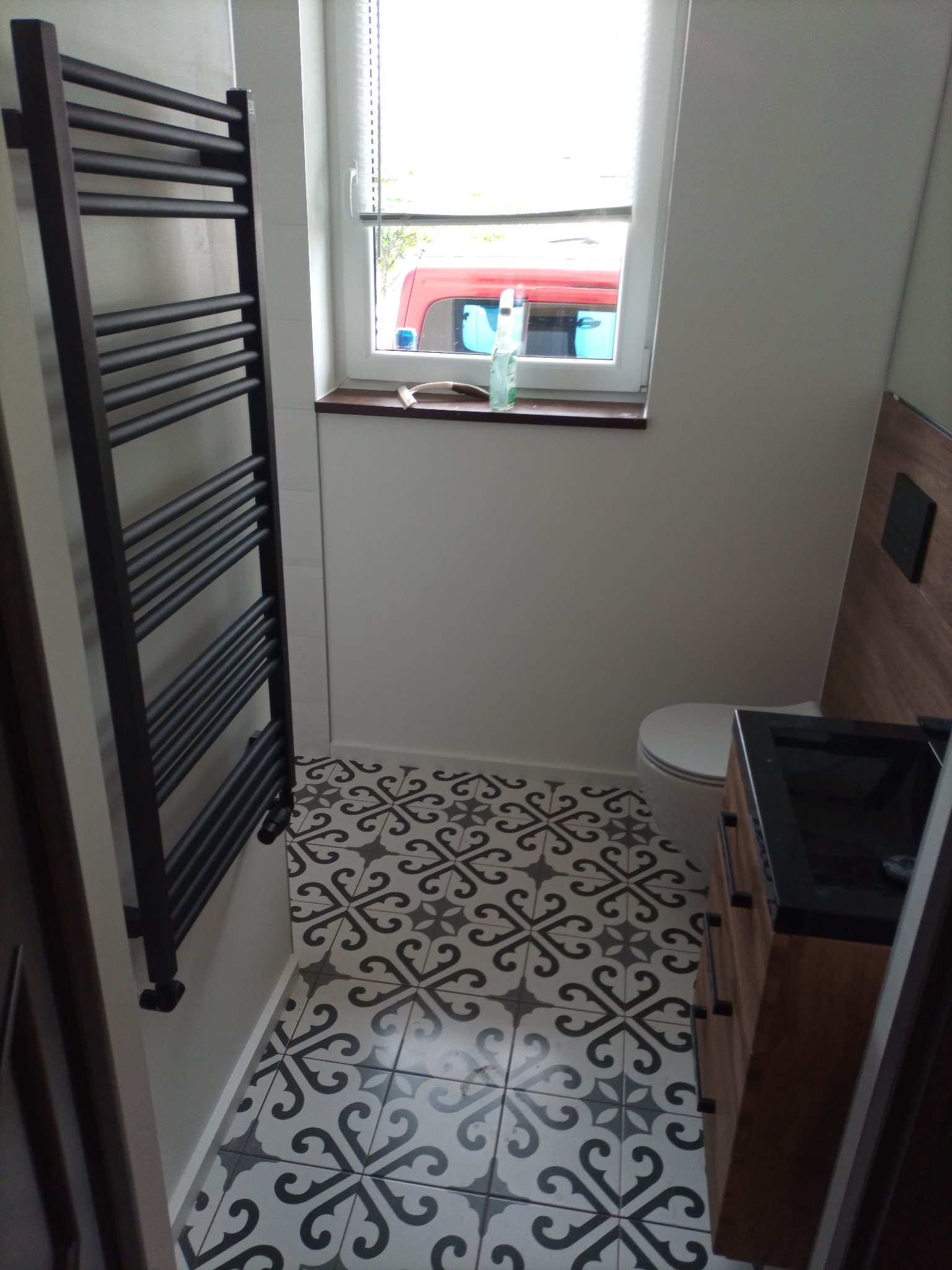Nowoczesna łazienka z czarno-białą wzorzystą podłogą, drewnianą szafką pod umywalką i grzejnikiem drabinkowym w kolorze antracytowym, widok na okno z butelką płynu do mycia.