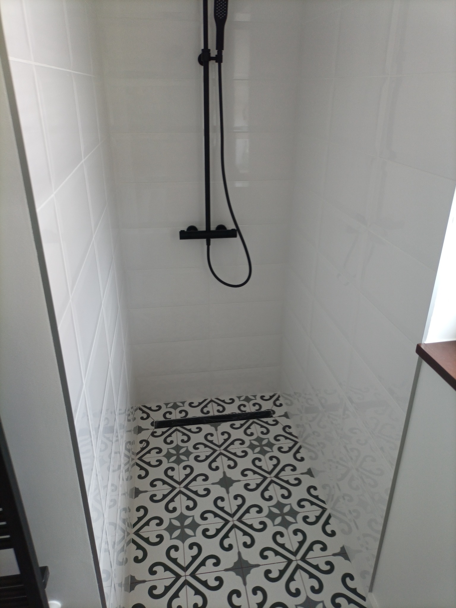 Wnętrze kabiny prysznicowej z białymi, błyszczącymi płytkami na ścianach i dekoracyjną, czarno-białą mozaiką na podłodze. Czarna armatura prysznicowa, w tym deszczownica i bateria, kontrastuje...