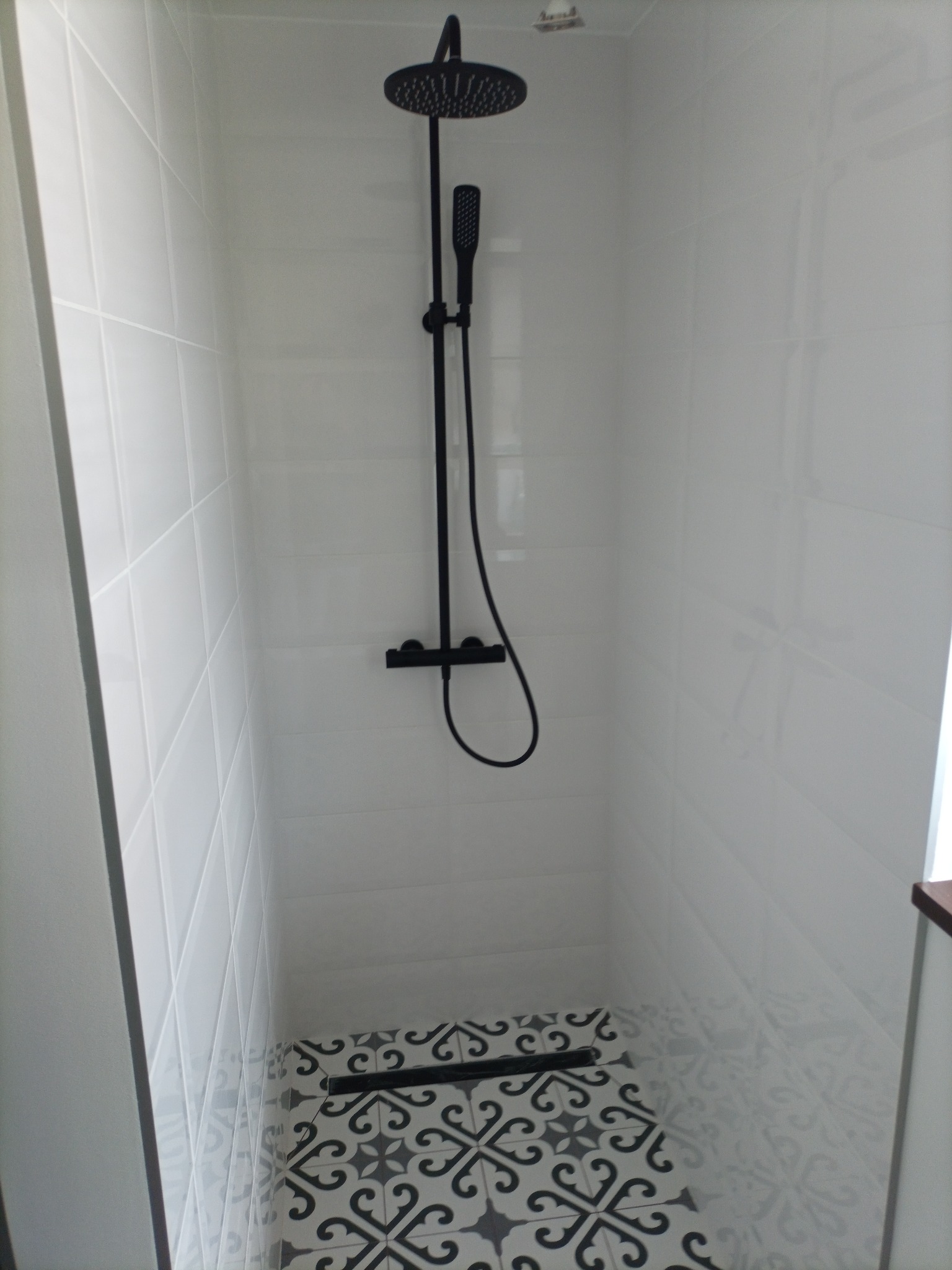 Nowoczesna kabina prysznicowa z czarną armaturą i wzorzystą podłogą w odcieniach szarości i bieli, wykończona białymi, błyszczącymi płytkami.