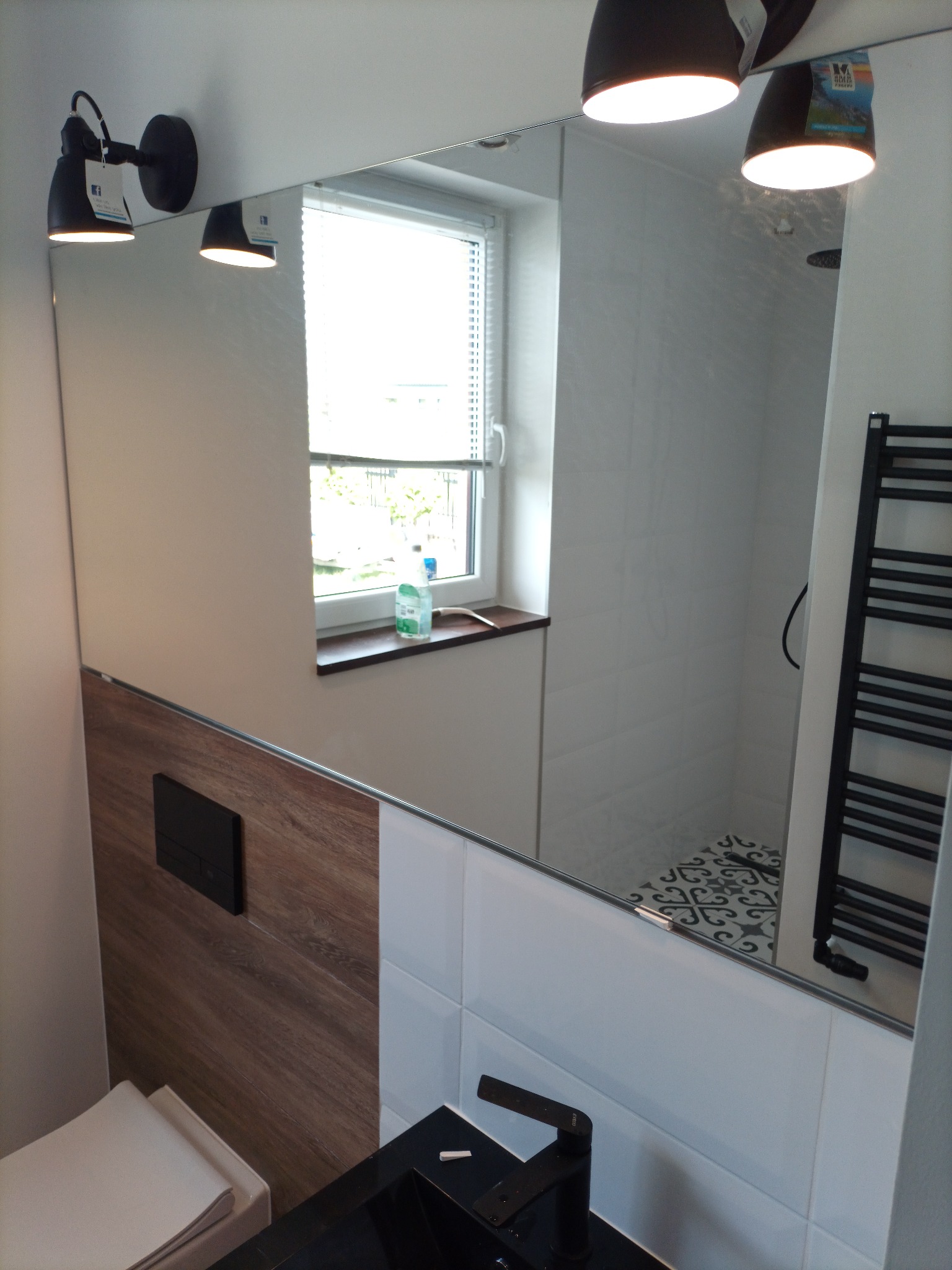 Nowoczesna łazienka z dużym lustrem, białymi płytkami i drewnianym akcentem, widok na kabinę prysznicową z dekoracyjnymi płytkami podłogowymi i grzejnik drabinkowy.