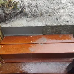 Schody drewniane z balustradą z okrąglaków, prowadzące do betonowego podwyższenia terenu.