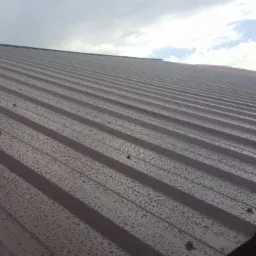 Ciemnobrązowe metalowe pokrycie dachowe z widocznymi kroplami deszczu, na tle pochmurnego nieba z prześwitami błękitu.