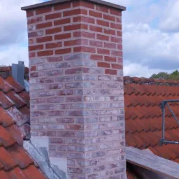 Ceglany komin na dachu z czerwonej dachówki, część komina odnowiona z jaśniejszymi cegłami, widoczne ślady świeżego spoiwa.
