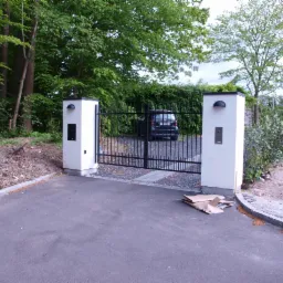 Czarna brama wjazdowa z białymi murowanymi słupkami, skrzynkami na listy i lampami, prowadząca na posesję z widocznym samochodem i zielenią w tle, na pierwszym planie leżą rozłożone kartony.