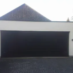 Biały garaż z czarną, segmentową bramą garażową, zwieńczony dwuspadowym dachem z szarej dachówki.