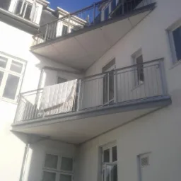 Elewacja budynku z balkonami z metalowymi balustradami, na jednym balkonie suszy się biała kołdra, widok z dołu pod kątem.