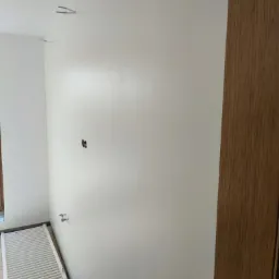 Świeżo pomalowana na biało ściana z widocznymi elementami instalacji elektrycznej i grzewczej, grzejnik na podłodze, widoczne ślady prac malarskich.