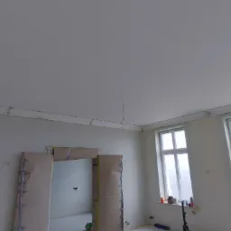 Pokój w trakcie przygotowań do malowania: zabezpieczone futryny kartonem, okna zakryte folią, sufit i ściany przygotowane do malowania, widoczna listwa dekoracyjna pod sufitem.