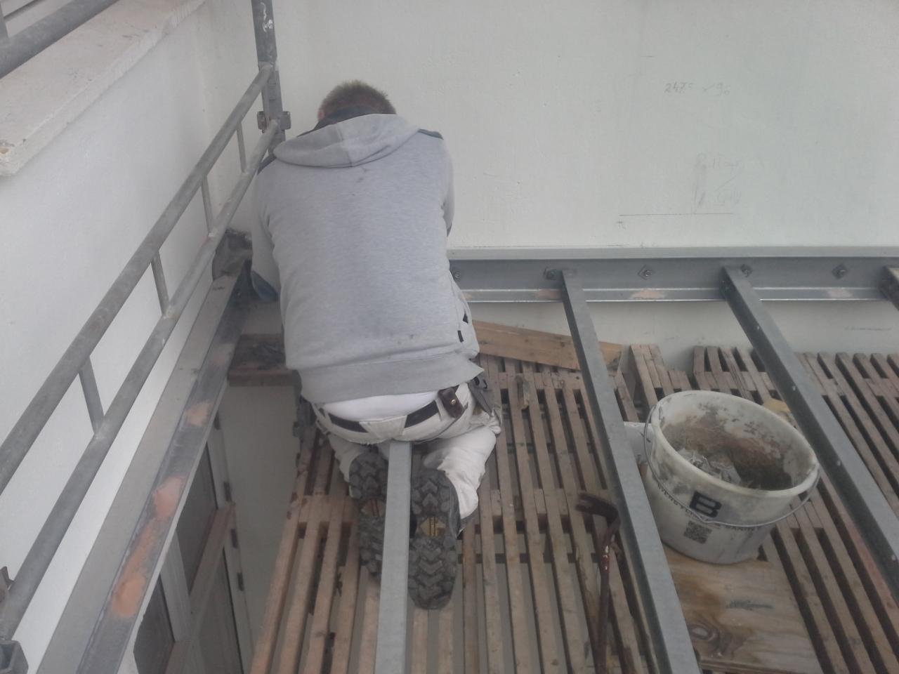 Pracownik w szarej bluzie i białych spodniach, klęczący na rusztowaniu podczas prac na wysokości, z widocznym wiadrem i narzędziami malarskimi, przed białą ścianą z zapiskami.