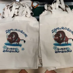 Stos beżowych toreb bawełnianych z niebieskim nadrukiem przedstawiającym stylizowanego żubra z napisem 'ZAMBROŻONE ŻUBRY ZAMBROW', ułożonych na stole.