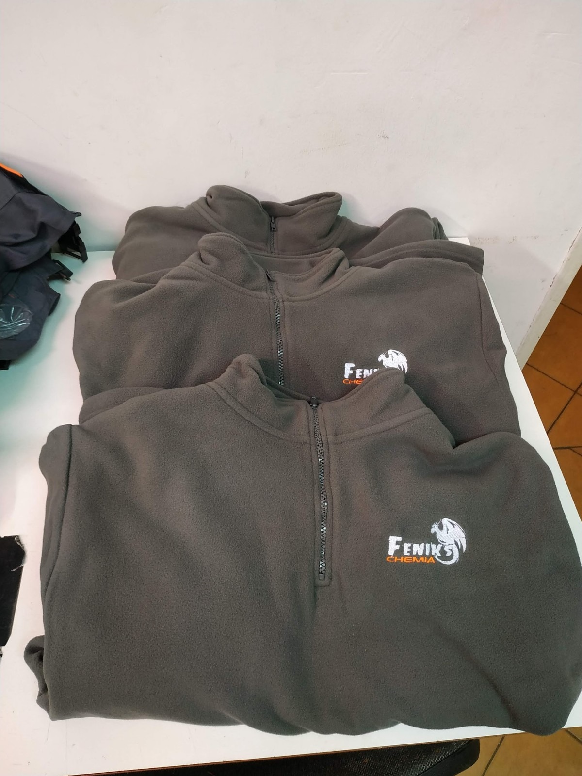 Trzy szare polarowe bluzy z zamkiem błyskawicznym i wyhaftowanym logo firmy 'Fenix Chemia' ułożone na białym blacie.