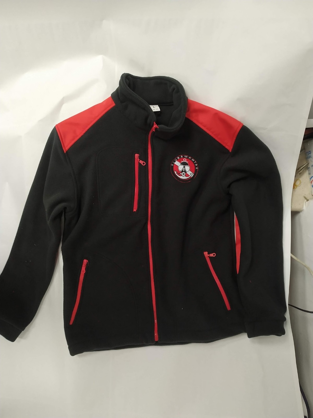 Czarna polarowa bluza z czerwonymi wstawkami na ramionach i czerwonymi zamkami błyskawicznymi, z haftowanym logo 'SCUBAMANIAK' na piersi, leżąca na białym tle.