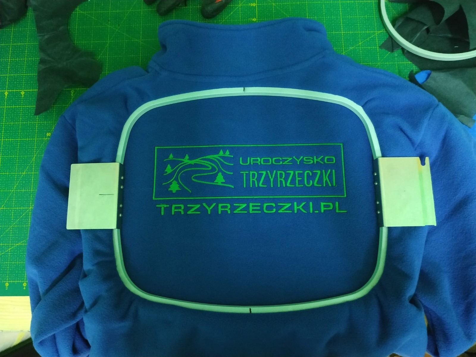 Niebieska bluza polarowa z zielonym haftowanym logo 'Uroczysko Trzyrzeczki.pl' w ramce do haftowania, widoczne fragmenty materiałów i narzędzi na zielonej macie do cięcia.