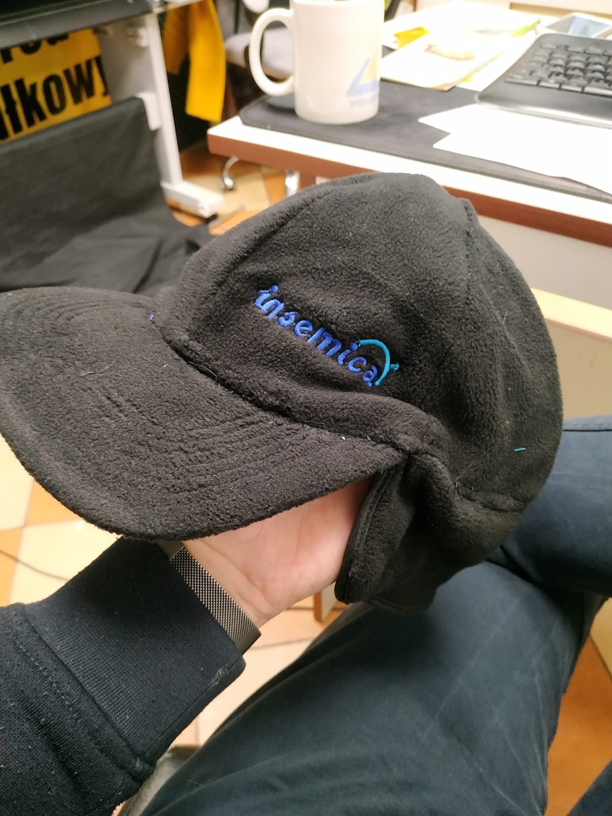 Czarna polarowa czapka z daszkiem z niebieskim haftowanym logo 'insemica' trzymana w dłoni, widoczny fragment rękawa i metalowy pasek zegarka, w tle biurko z kubkiem i klawiaturą.