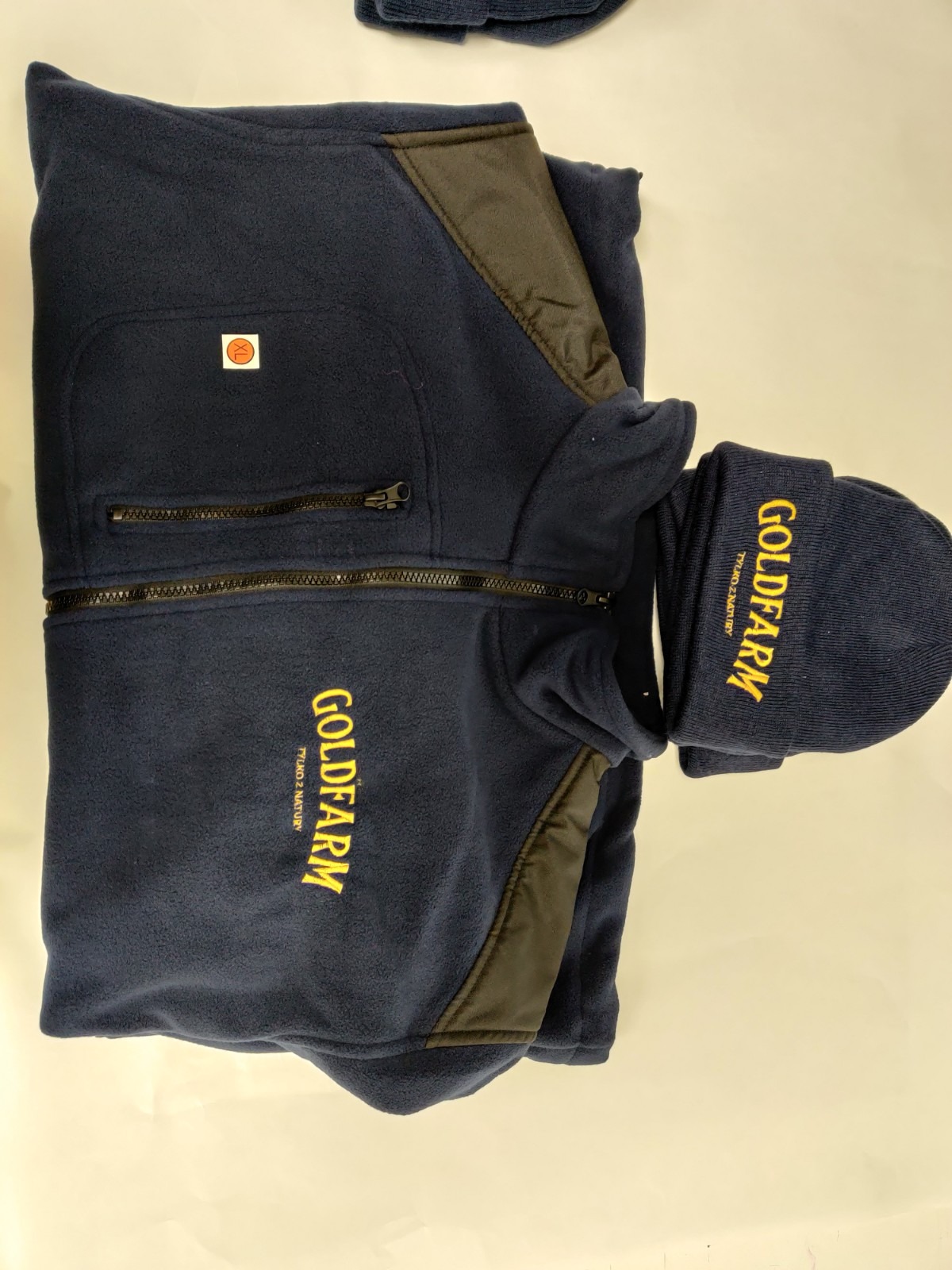 Granatowa bluza polarowa rozpinana z czarnymi wstawkami i żółtym haftem logo 'GOLDFARM TYLKO Z NATURY' oraz pasująca czapka z takim samym haftem, leżące na jasnym tle.