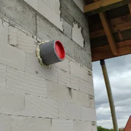 Szary przewód wentylacyjny z czerwoną zaślepką wystaje ze ściany budynku z betonu komórkowego, uszczelniony pianką montażową. W tle fragment dachu i pochmurne niebo.