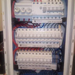 Wnętrze skrzynki rozdzielczej z bezpiecznikami i okablowaniem w kolorach niebieskim i czerwonym, zamontowanej w białej obudowie.