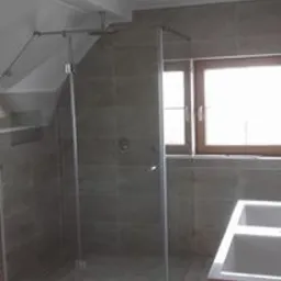 Szara kabina prysznicowa z przezroczystymi szklanymi drzwiami w łazience ze skośnym sufitem, wykończona płytkami w odcieniach szarości i beżu, widoczne okno z drewnianą ramą.