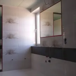 Biało-szara łazienka z płytkami w kwiaty, widoczne przygotowane punkty instalacyjne i lustro z czerwoną ramą, oraz fragment okna.