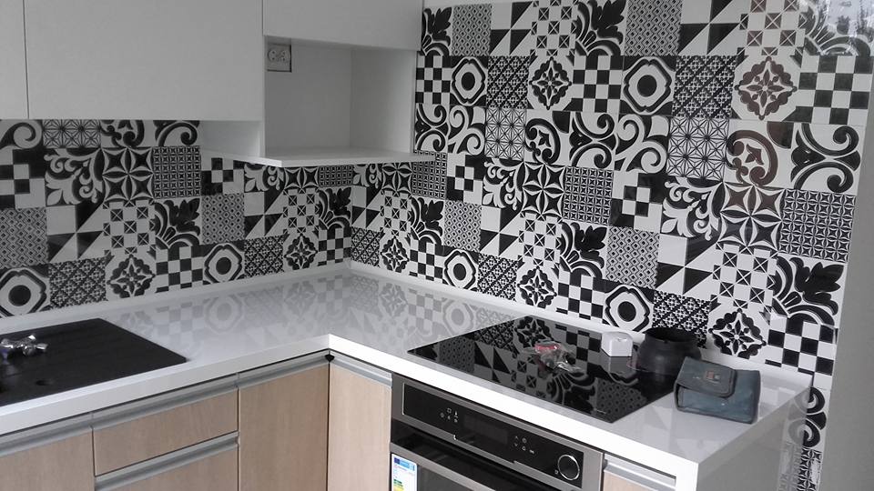 Kuchnia z czarno-białymi płytkami patchwork na ścianie nad blatem i płytą indukcyjną, widoczne szafki z jasnego drewna i białe górne szafki.