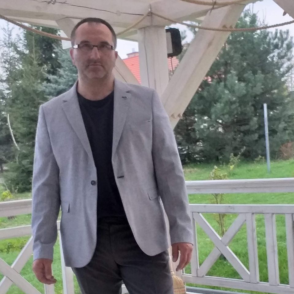 Mężczyzna w okularach i szarej marynarce stoi na drewnianym tarasie altany na tle zieleni.