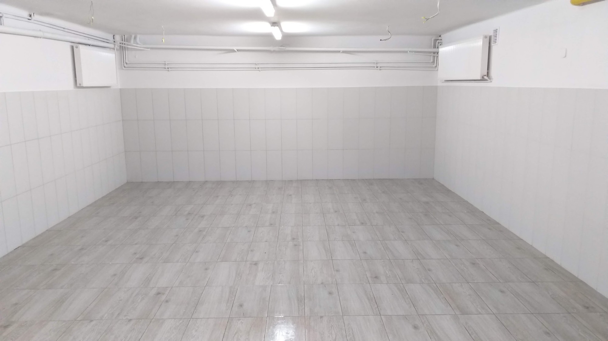 Puste pomieszczenie z wyłożoną jasną, imitującą drewno, podłogą z płytek oraz białymi płytkami ściennymi do połowy wysokości, powyżej pomalowane na biało, widoczne rury i grzejniki.