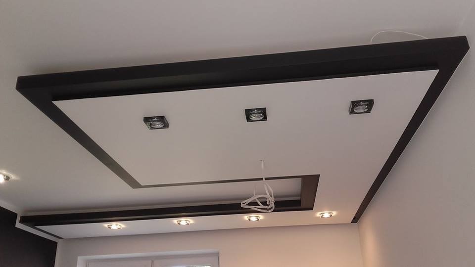 Biały sufit podwieszany z czarną obwódką, oświetlony kwadratowymi lampami, z widocznymi kablami w jednym z otworów.