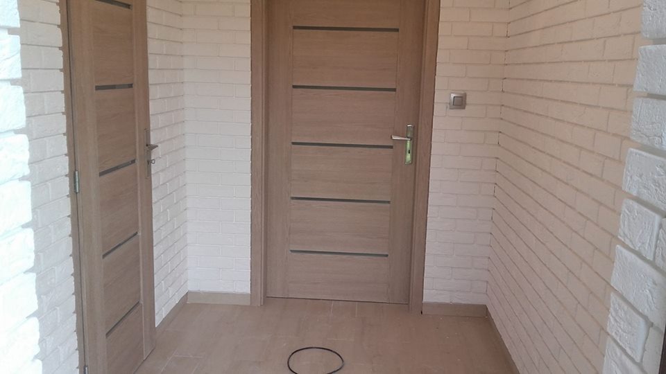 Dwie drewniane drzwi z poziomymi, ciemnymi wstawkami, na tle ściany z białej cegły, wewnątrz pomieszczenia z jasną podłogą z płytek.