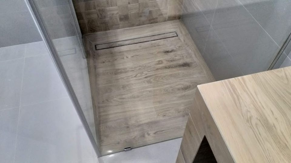 Kabina prysznicowa walk-in z odpływem liniowym i płytkami imitującymi drewno na podłodze, jasnoszare płytki ścienne, częściowo widoczna szafka pod umywalkę z frontami w tym samym odcieniu drewna.