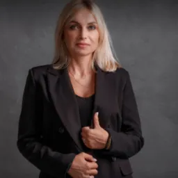 Portret kobiety w czarnej marynarce na tle szarej ściany z widocznym logo firmy w prawym górnym rogu.