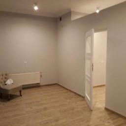 Kamil Barma Zakład Usług Ogólnobudowlanych - Pusty pokój po remoncie z jasnoszarą ścianą, panelami podłogowymi i otwartymi białymi drzwiami, z beżowym fotelem i białymi kwiatami. Widoczne dwa reflektory sufitowe.