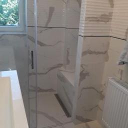 Kamil Barma Zakład Usług Ogólnobudowlanych - Kabina prysznicowa z białymi płytkami imitującymi marmur, z widocznym odpływem liniowym i fragmentem okna z zielenią na zewnątrz.