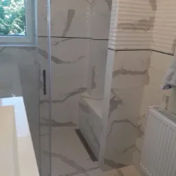 Kabina prysznicowa z białymi płytkami imitującymi marmur, z widocznym odpływem liniowym i fragmentem okna z zielenią na zewnątrz.