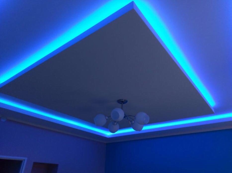 Podwieszany sufit z oświetleniem LED w kolorze niebieskim, widoczny z perspektywy narożnika pokoju, z żyrandolem z mlecznymi kulami w centralnej części.