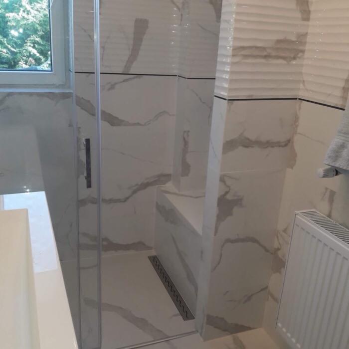 Kabina prysznicowa z białymi płytkami imitującymi marmur, z widocznym odpływem liniowym i fragmentem okna z zielenią na zewnątrz.