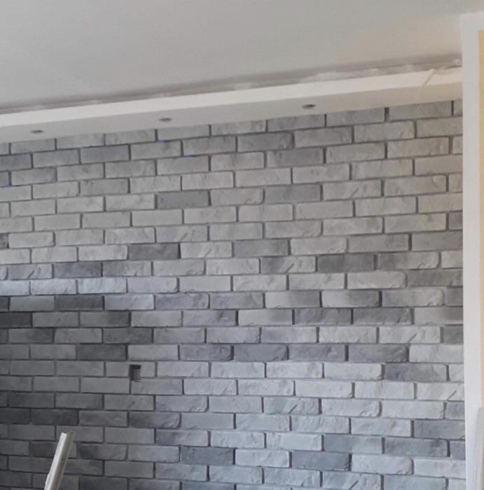 Ściana wyłożona szarą cegłą dekoracyjną, oświetlona punktowymi lampami w suficie podwieszanym. Widoczne niedociągnięcia przy montażu.