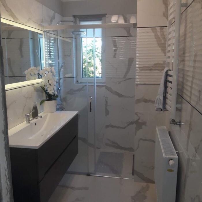 Nowoczesna łazienka z kabiną prysznicową, umywalką w szafce i grzejnikiem drabinkowym, wykończona płytkami imitującymi marmur z szarymi żyłami.