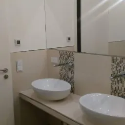 Nowoczesna łazienka z dwiema białymi, owalnymi umywalkami nablatowymi, osadzonymi na beżowym blacie. Ściany wykończone beżowymi płytkami z dekoracyjnym pasem mozaiki. Duże lustro z widocznym...