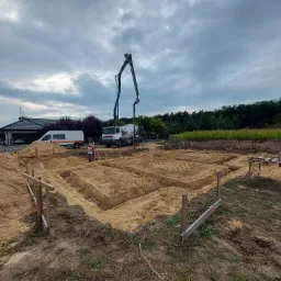 Przygotowanie terenu pod fundamenty domu jednorodzinnego: wykopane rowy zbrojeniowe wypełnione piaskiem, na tle betoniarka z pompą oraz pracownik w kasku.