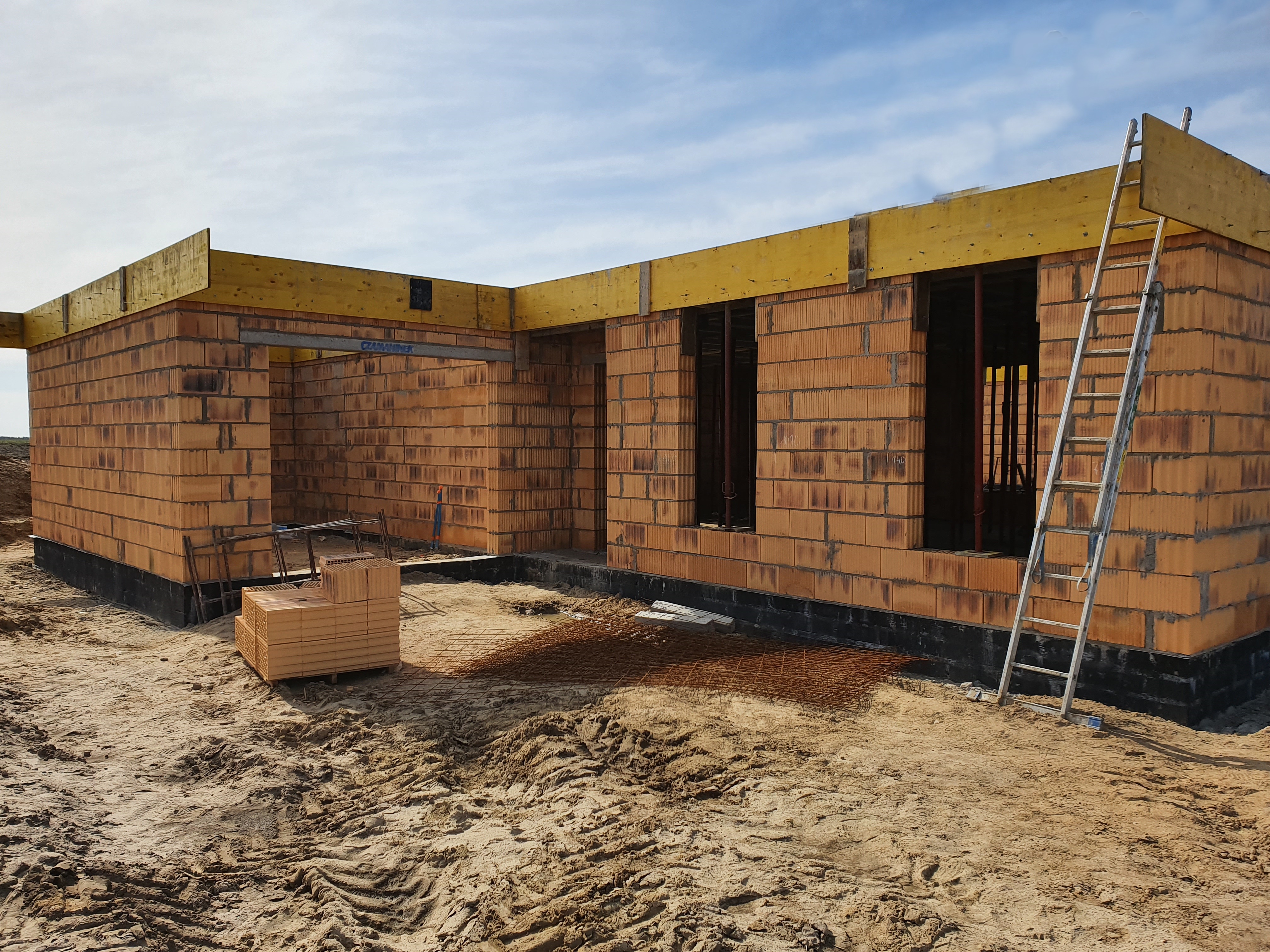 Wznoszenie ścian parterowego domu z pustaków ceramicznych, widoczna drabina oparta o ścianę, żółte drewniane nadproża i stos pustaków obok budynku.