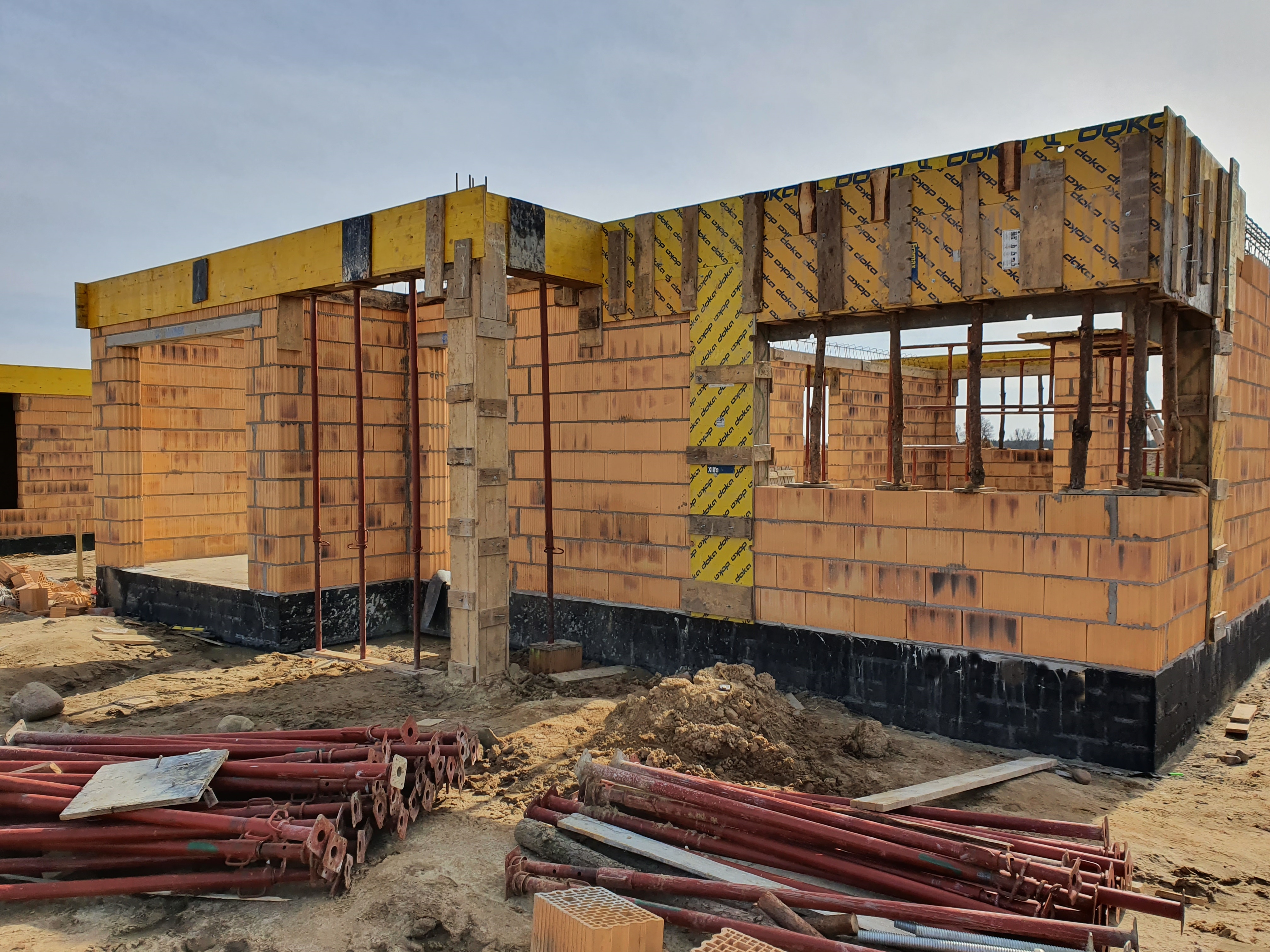 Budowa domu w trakcie realizacji: widoczne ściany z pustaków ceramicznych, żółte szalunki stropowe Doka, podpory budowlane w kolorze czerwonym, czarna izolacja przeciwwilgociowa fundamentów.