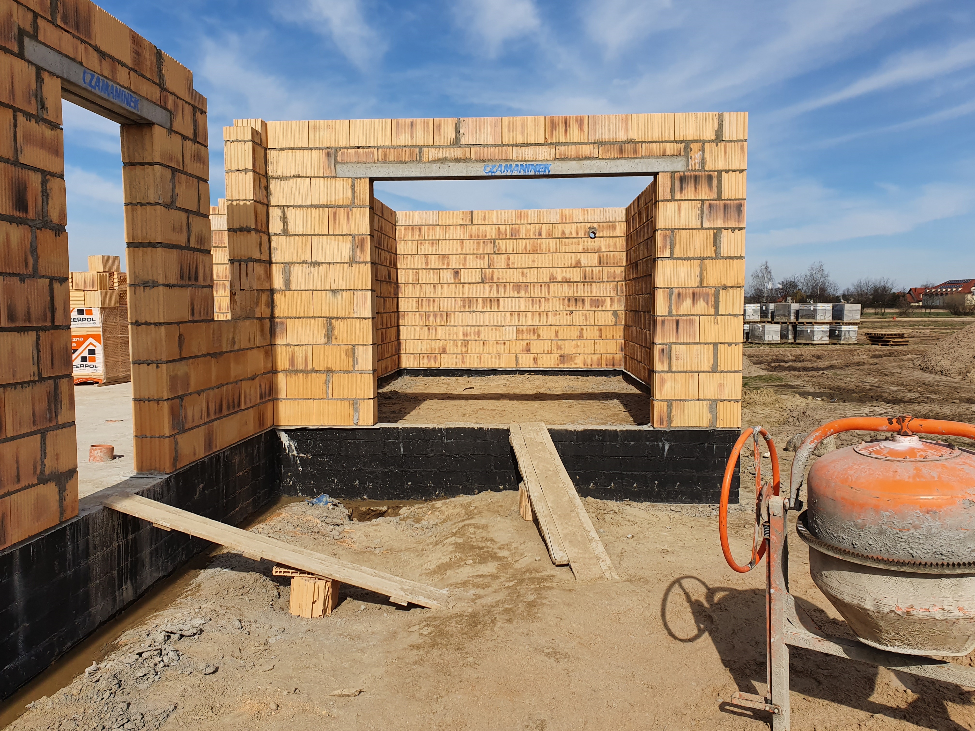 Wznoszenie ścian budynku z pustaków ceramicznych, widoczny betonowy nadproże z niebieskim napisem, na pierwszym planie pomarańczowa betoniarka, w tle palety z pustakami i błękitne niebo.