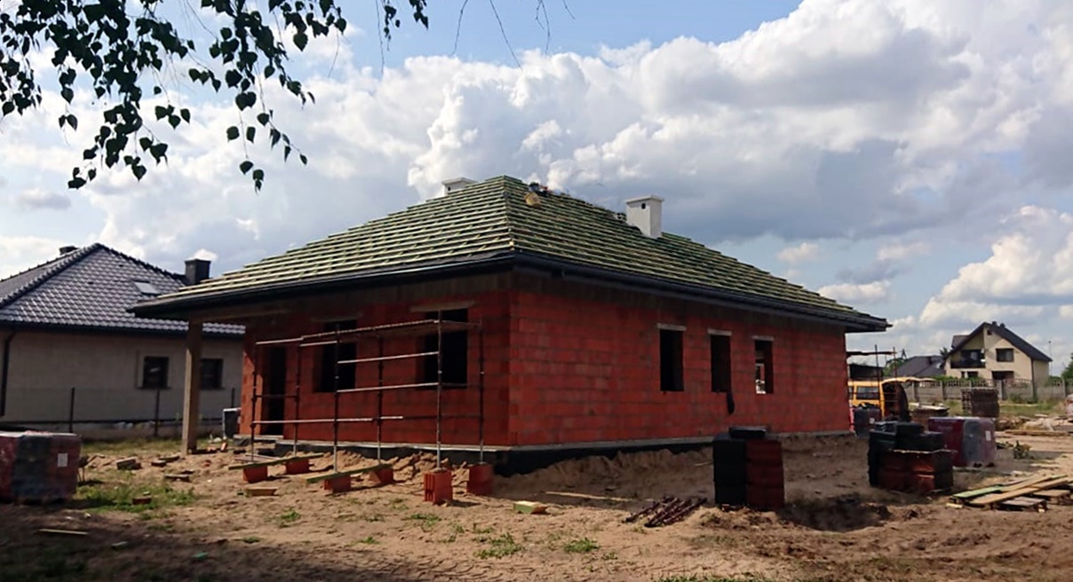 Surowy, jednokondygnacyjny budynek z czerwonej cegły z zielonym dachem, częściowo zadaszony, z rusztowaniem przed otworami okiennymi, na tle pochmurnego nieba i innych domów w oddali.