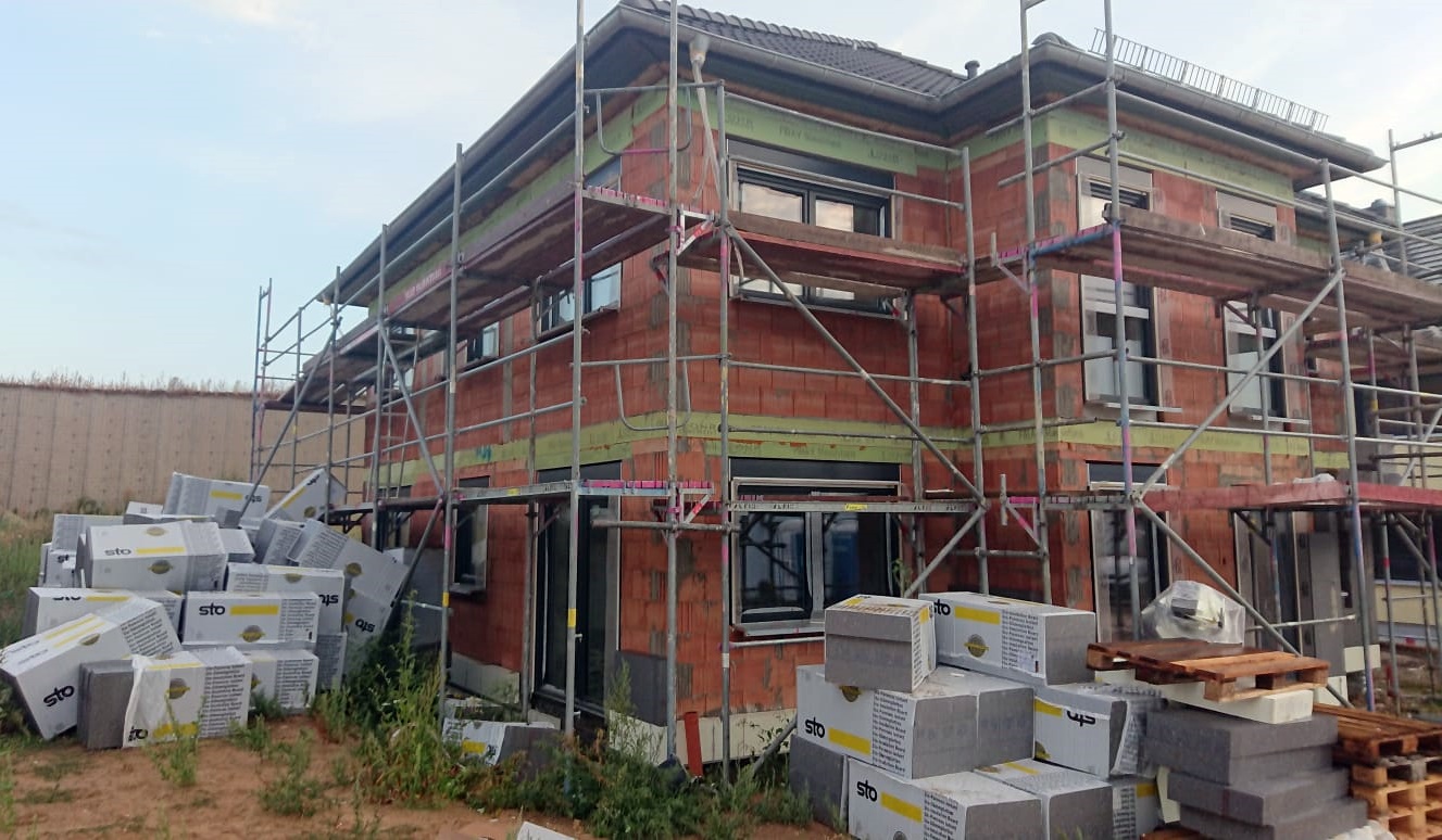 Wznoszony dwukondygnacyjny budynek z czerwonej cegły, z widocznymi rusztowaniami, oknami w czarnych ramach i ociepleniem, z paletami materiałów budowlanych STO na pierwszym planie.