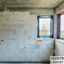 Surowy stan wnętrza z widocznymi przewodami elektrycznymi poprowadzonymi po ścianach z bloczków, okablowanie w peszlach przy podłodze, okna z folią ochronną, ściany z notatkami i oznaczeniami.